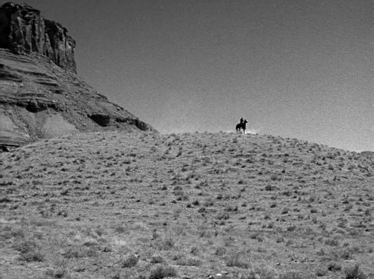 Rio Grande (1950) movie still: wide — A vast, dry desert landscape with a steep rocky hill and sparse vegetation. A single rider on a horse stands…