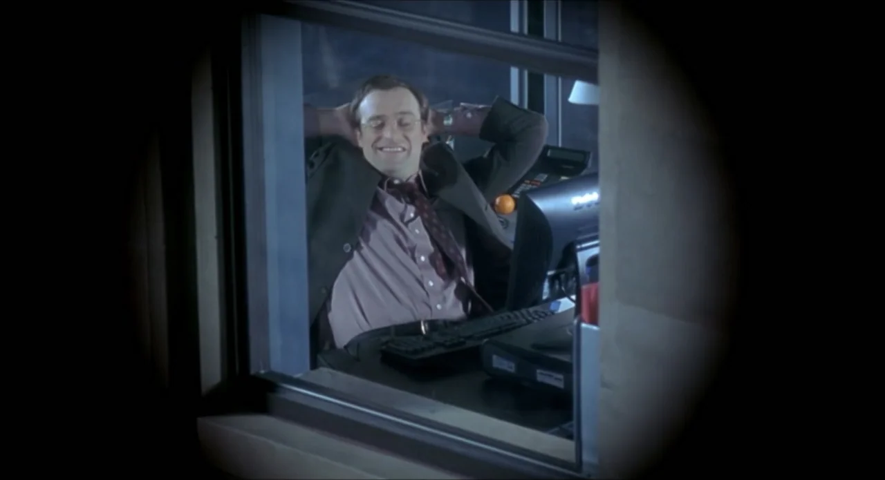 Foolproof (2003) movie still: medium — An office interior at night framed through a window frame with vignette borders. A man in business attire…