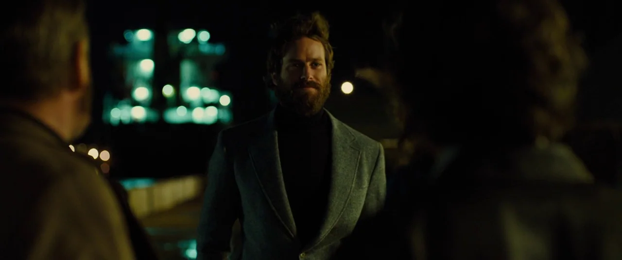 Free Fire (2016) movie still: medium — A man stands outdoors at night with two other people partially framing him. A blurred industrial structure…
