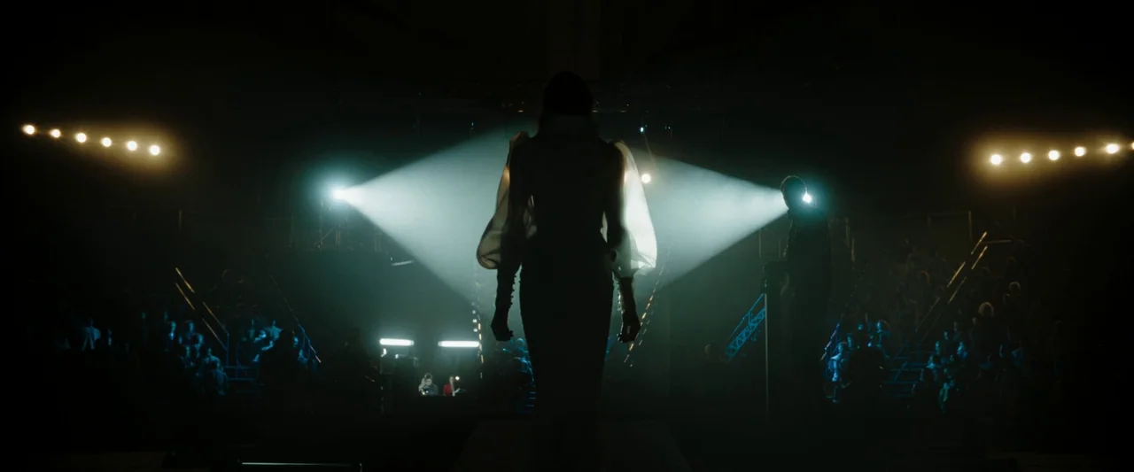 Boy Kills World (2023) movie still: fullbody — A high-fashion runway show takes place in a dark, dimly lit industrial space. A model wearing a dress…
