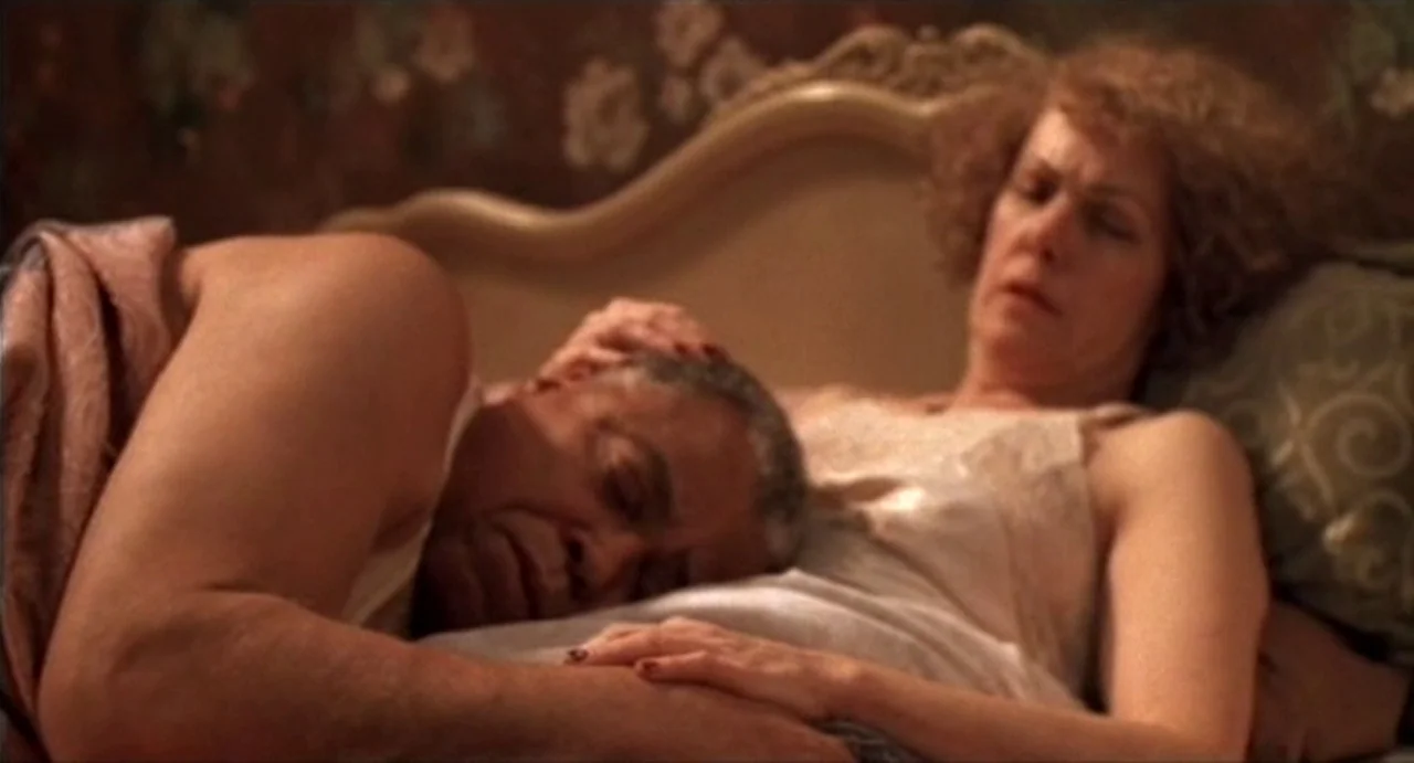 The Annihilation of Fish (1999) movie still: medium — A couple lies in a bed against a patterned floral wallpaper and upholstered headboard. The man…