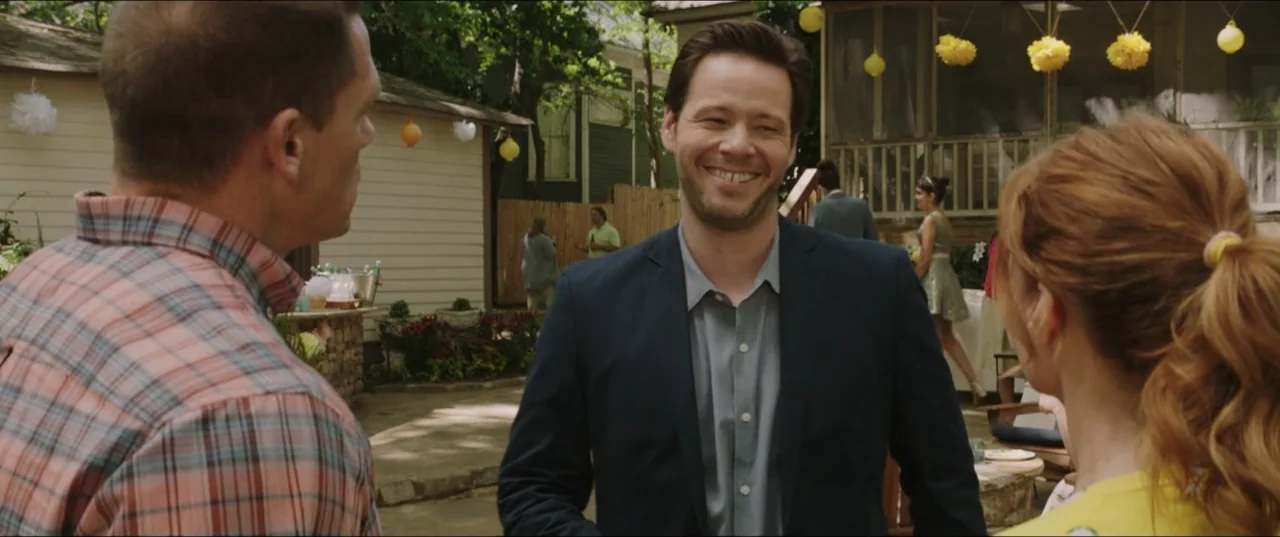 Blockers (2018) movie still: medium — A backyard party features people gathering in the background near a light-colored building. A man in a dark…