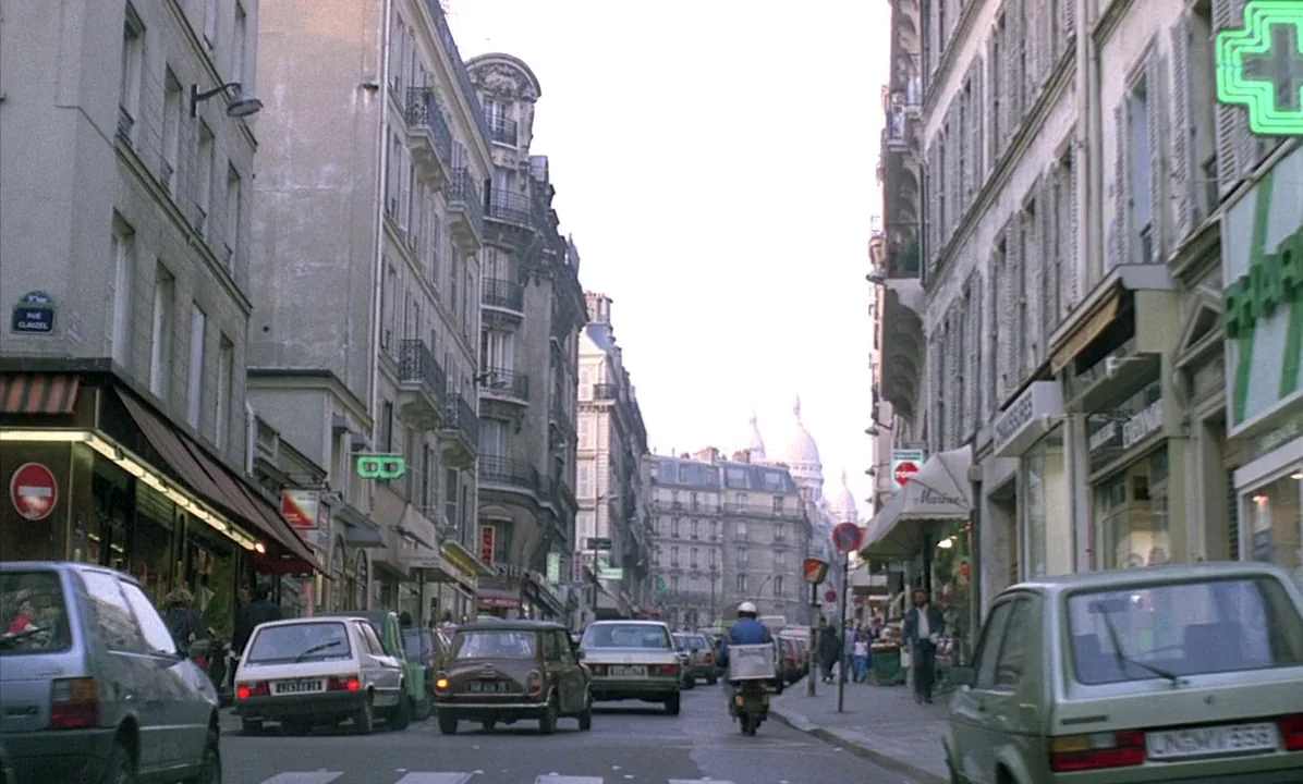 A Tale of Springtime (1990) movie still: establishing — A narrow European city street is lined with historic multi-story stone buildings and…