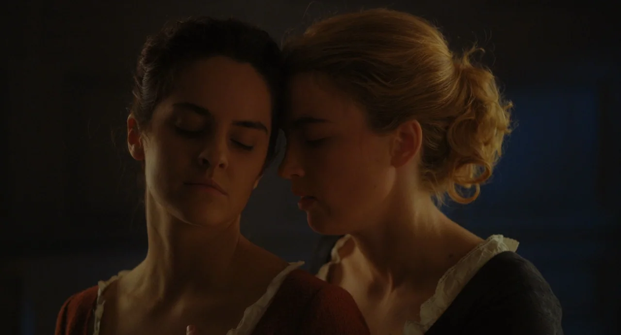Portrait of a Lady on Fire movie still: closeup — A dimly lit room provides a background for two women standing together. They rest their heads…