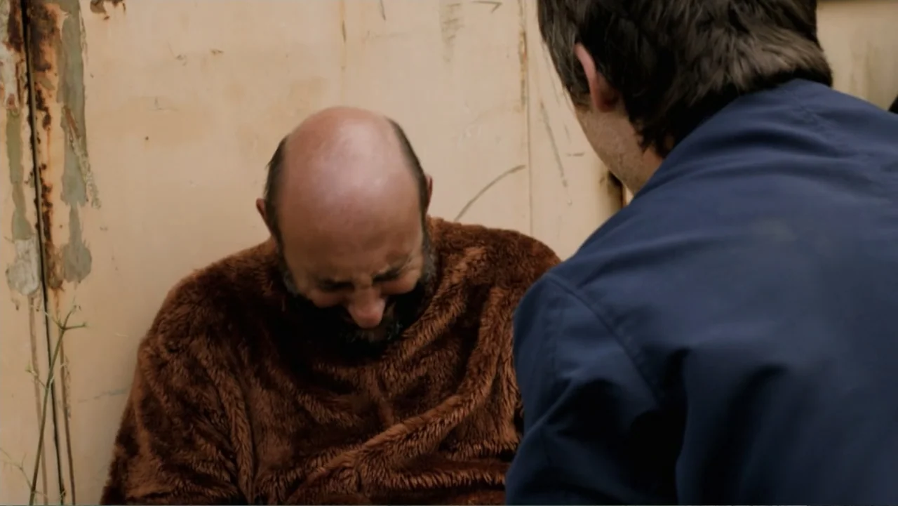 A Boyfriend for My Wife (2008) movie still: medium — A man in a brown fleece sweater sits slumped against a weathered metal wall. A second person in…