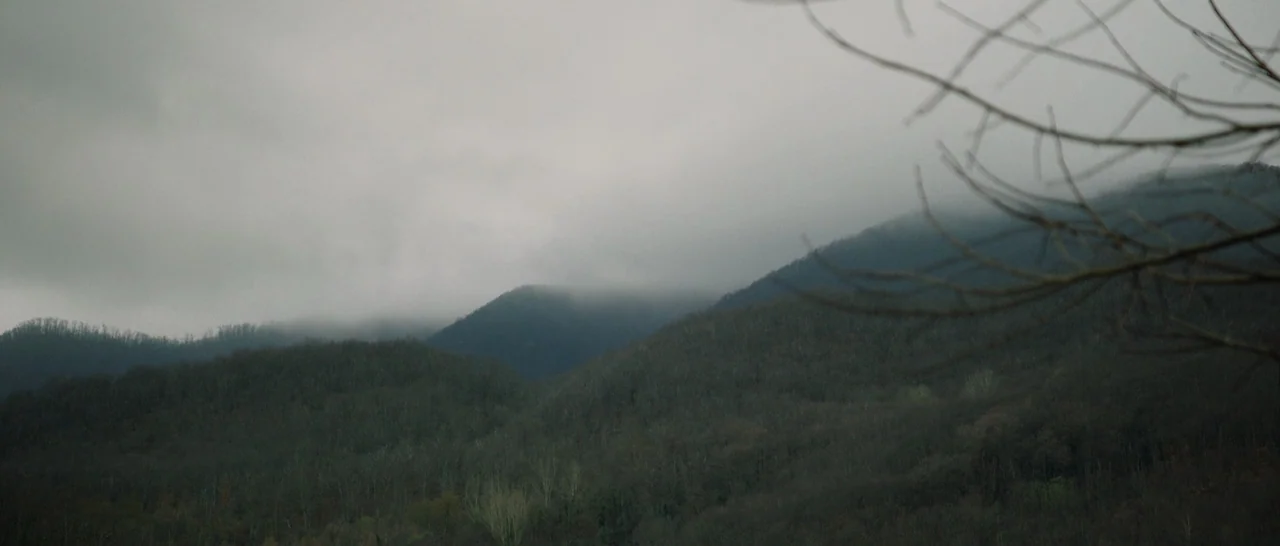 The Little Hours (2017) movie still: establishing — A dense, treelined mountain range rises beneath a heavy, overcast grey sky. Low-hanging clouds…