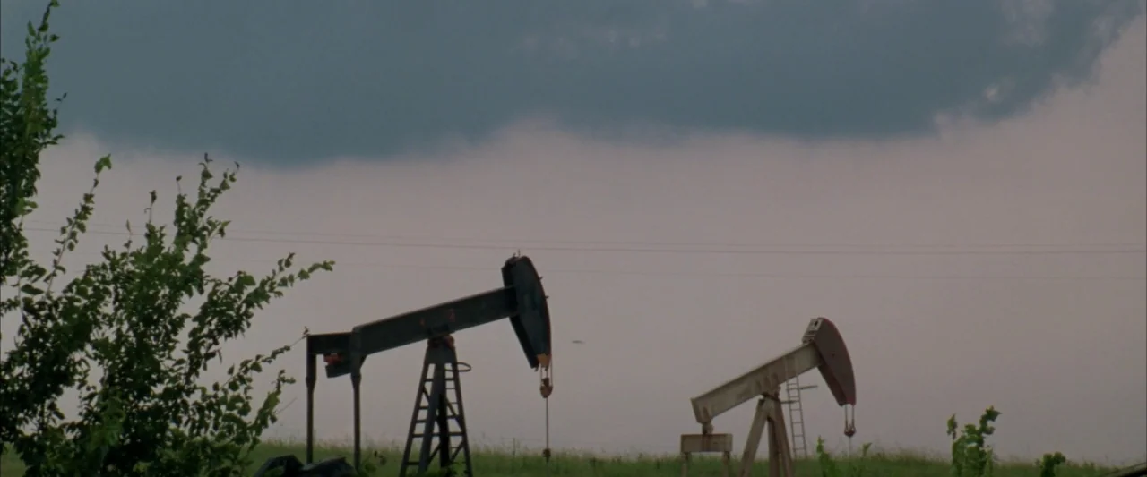 Twister (1996) movie still: wide — Two industrial oil pumpjacks stand in an open, grassy landscape under overcast skies. Green foliage frames the…