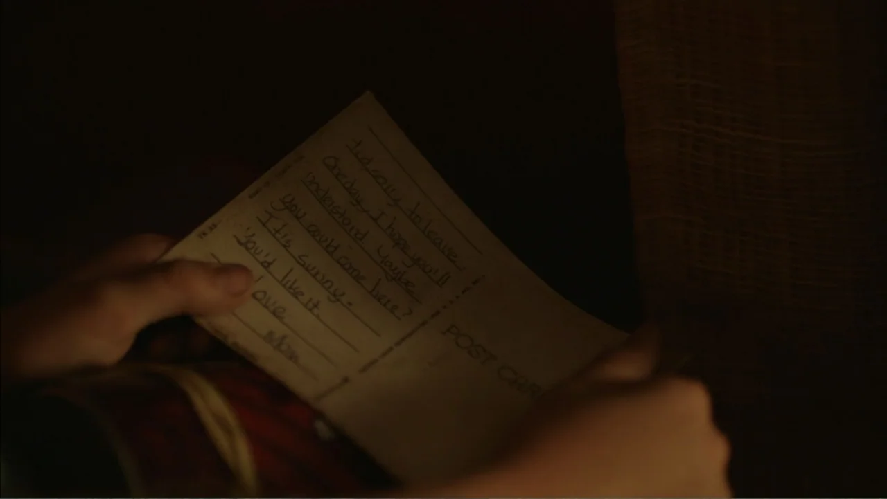 Boy (2010) movie still: closeup — A close-up shot focuses on hands holding a vintage postcard with handwritten ink text. The background is a dark,…
