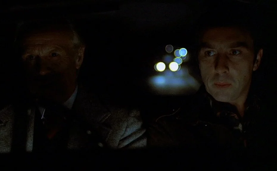 To The Devil a Daughter (1976) movie still: medium — Two men sit side-by-side in the front seat of a dark car at night. Outside the windshield, the…