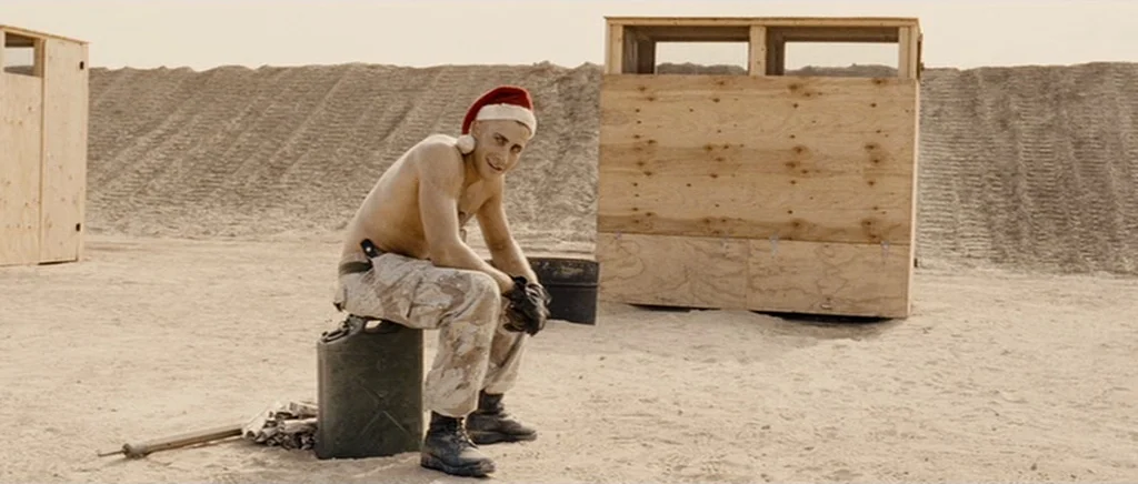Jarhead movie still: fullbody — A flat desert training site features plywood structures and deep tire tracks in sand. A shirtless man sits on a dark…