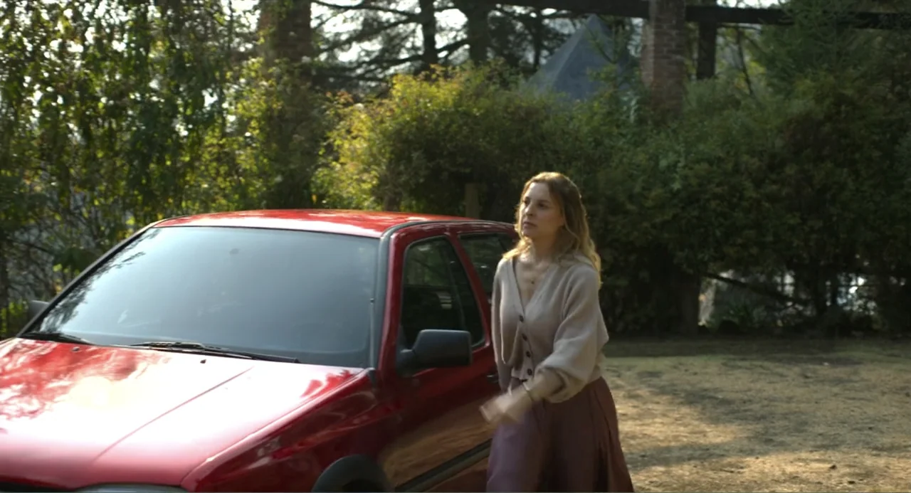 Heartbeat (2008) movie still: fullbody — An outdoor paved area sits beside a tall brick structure partially obscured by green trees. A woman in a…