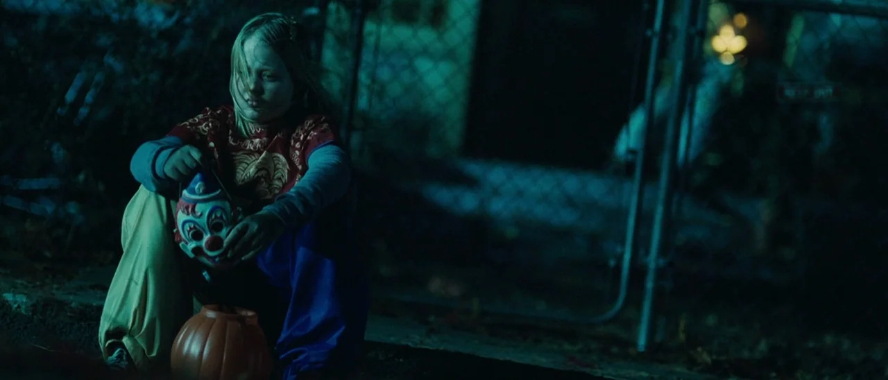 Halloween (2007) movie still: medium — A child sits on a dark residential curb next to a chain-link fence at night. She holds a plastic clown mask…