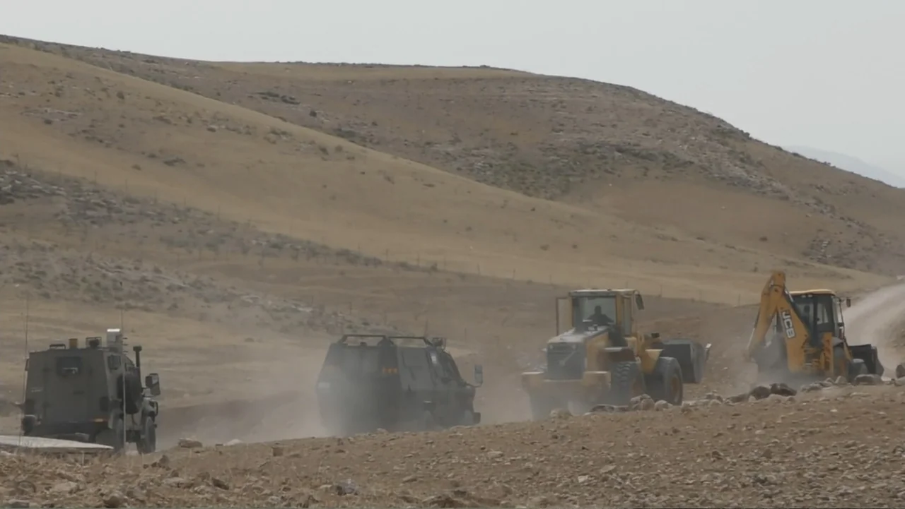 No Other Land (2024) movie still: wide — A dry hilly landscape features a dirt road with several heavy vehicles. A military-style truck follows a…