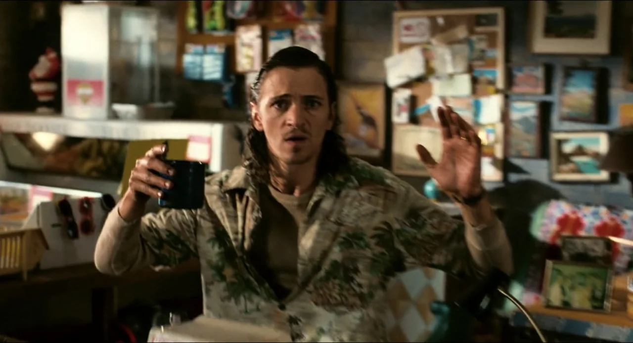 From Dusk Till Dawn (1996) movie still: medium — A room filled with various framed photographs and cluttered shelves on the walls. A young man sits…