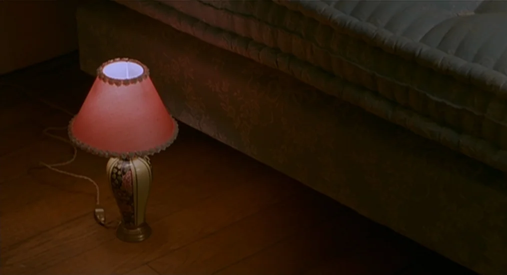 Vendredi Soir (2002) movie still: closeup — A vintage table lamp with a patterned base and fabric shade sits on hardwood. The lamp illuminates the…