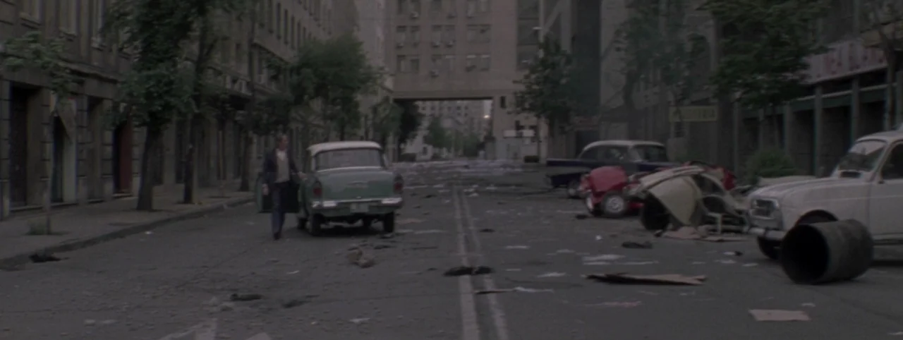 Post Mortem (2010) movie still: wide — An empty urban street scattered with debris is flanked by tall, older concrete buildings. A lone man exits a…