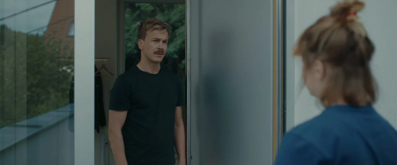 Peacock (2010) movie still: medium — A man stands in the doorway of a modern building looking toward a woman. The interior space features a glass…