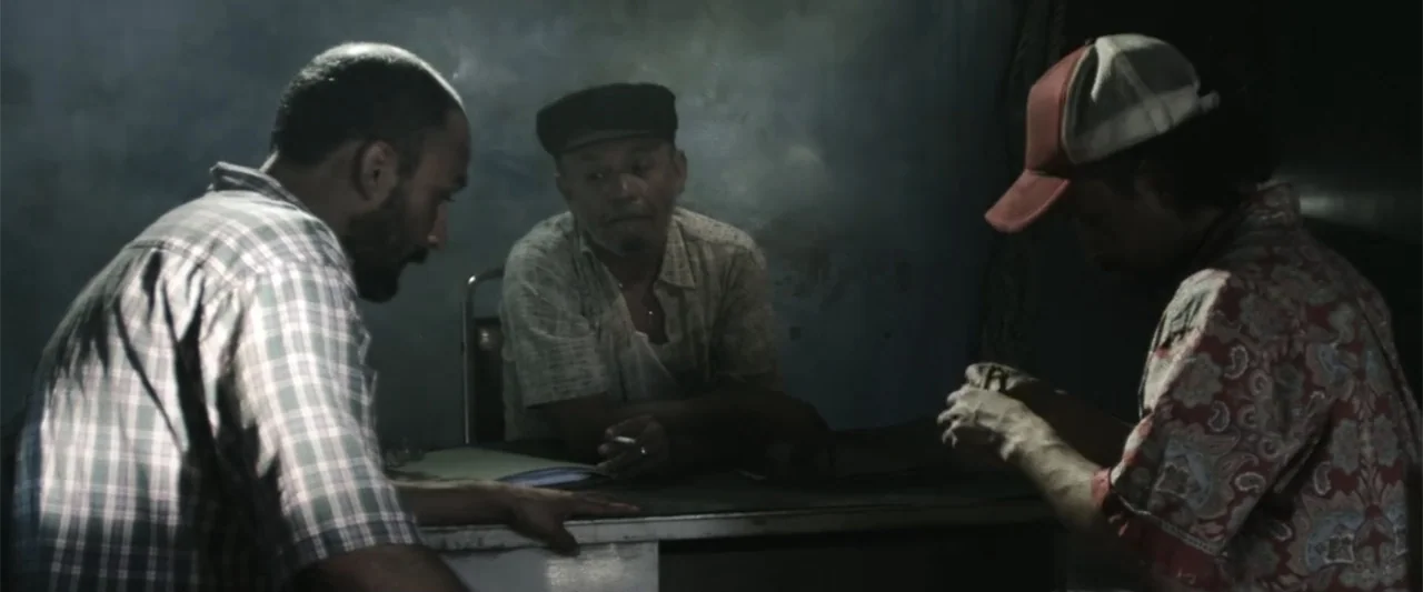 At Dawn (2013) movie still: medium — A dimly lit room with blue-tinted walls and a desk holding papers. Three men sit closely together at the desk…