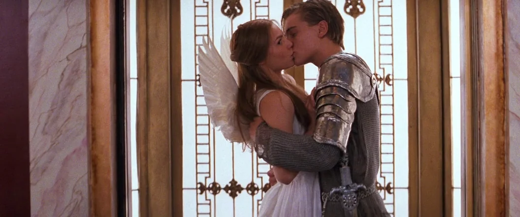Romeo + Juliet (1996) movie still: medium — A pair of teenagers in costumes stand before an ornate gilded metal screen doorway. A woman dressed as…