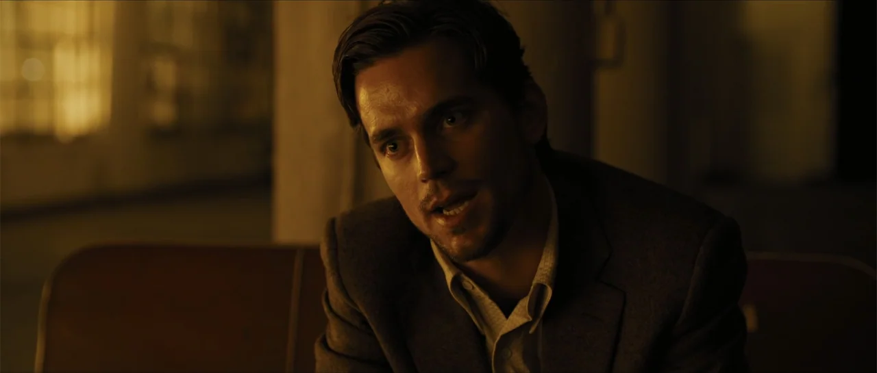 In Time (2011) movie still: medium — A dimly lit room with muted golden walls and soft shadows. A man in a blazer sits while engaged in conversation.