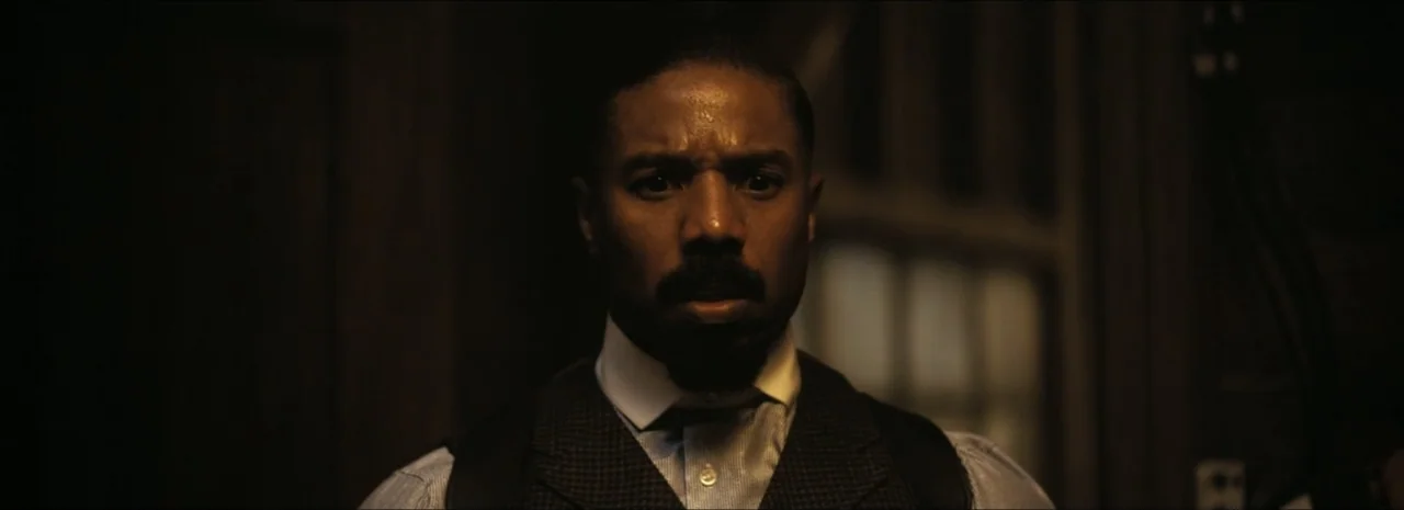 Sinners (2025) movie still: closeup — A man stands in a dark room with a single window behind him. He gazes directly at the camera with a wide-eyed,…