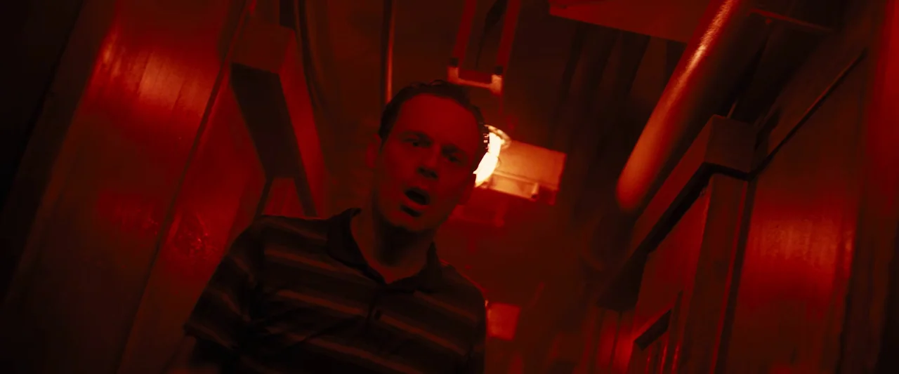 Black Sea (2014) movie still: medium — A narrow metallic corridor is illuminated by a piercing red emergency light. A man stands centrally, staring…