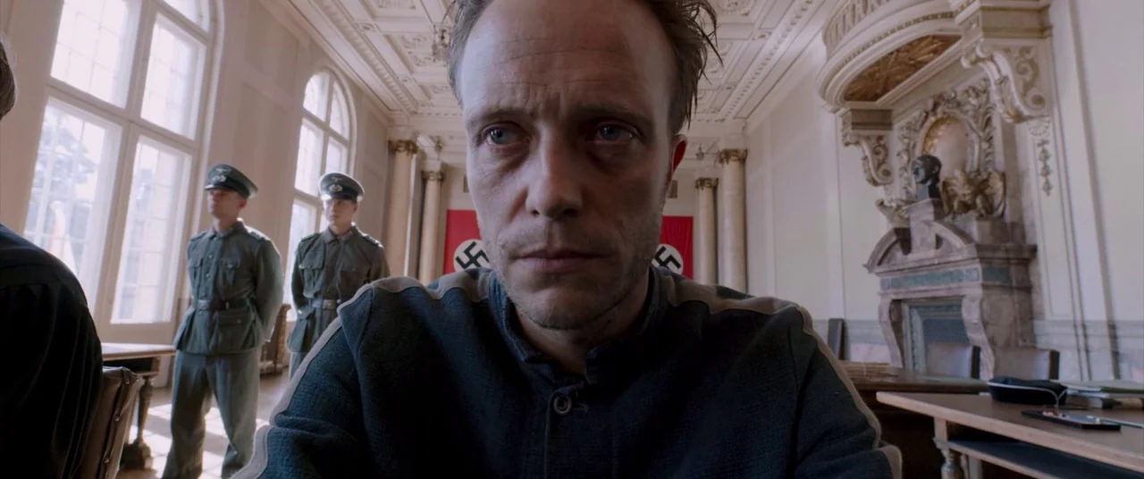 A Hidden Life (2019) movie still: closeup — A high-ceilinged room with large windows and ornate architectural molding displays a red flag. A man…