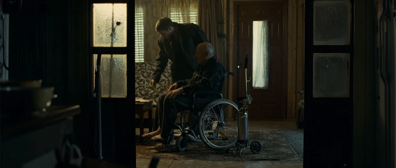 Blood Creek (2009) movie still: medium — A cluttered living room features wood-paneled walls and a worn rug on the floor. A younger man stands…