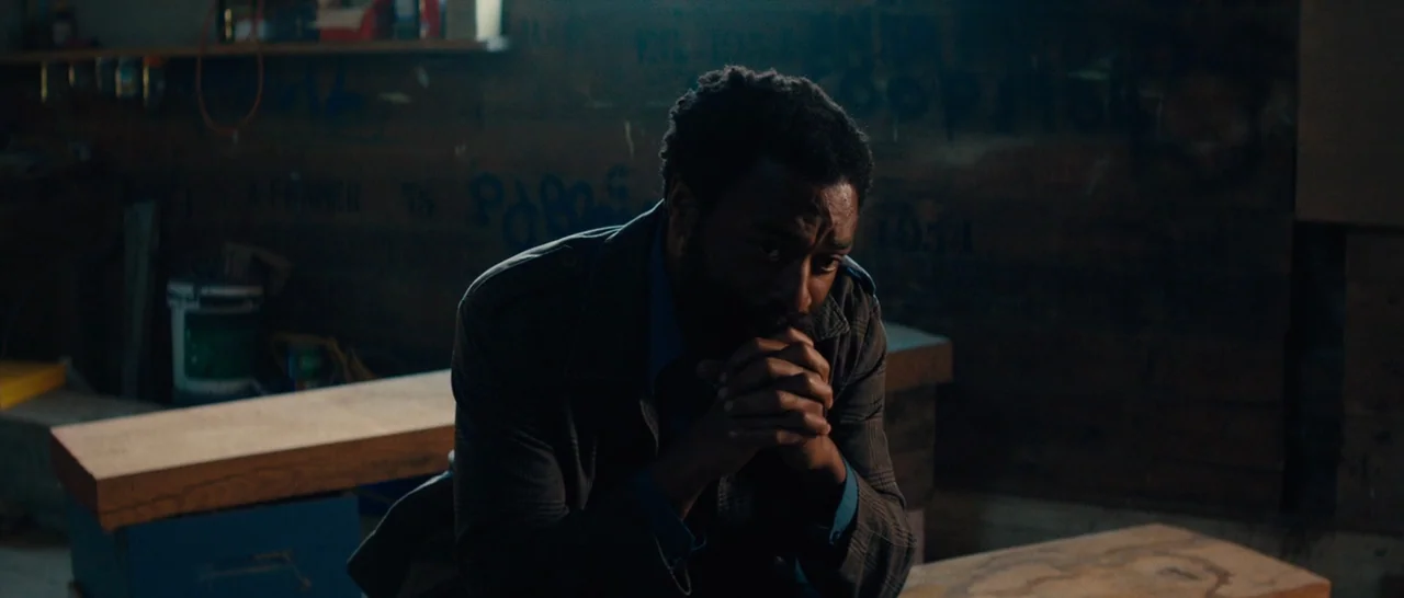 Z for Zachariah (2015) movie still: medium — A cluttered workshop with wooden benches and wall markings under dim light. A man sits alone with his…
