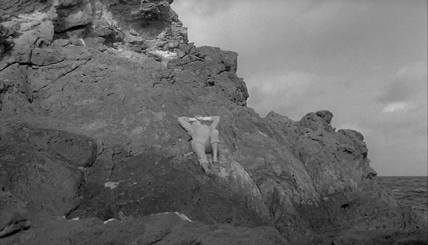 L’Avventura (1960) movie still: wide — A rugged rocky coastline with uneven dark stone faces the open sea. A man rests on his back on a flat section…