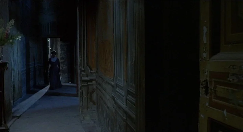 Lisa and The Devil (1973) movie still: wide — A dimly lit narrow hallway with wood paneling and aged wallpaper finish. A woman stands in a distant…