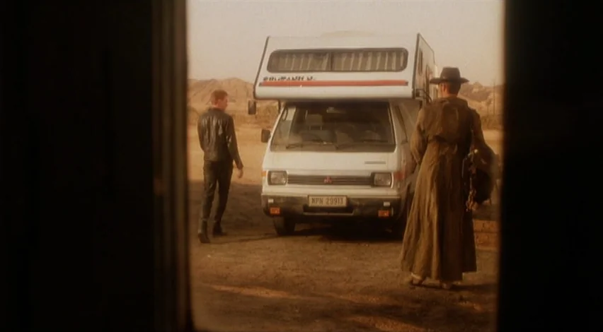 Dust Devil (1992) movie still: wide — A dry desert landscape features a white camper van parked in the center. Two men walk toward the vehicle from…