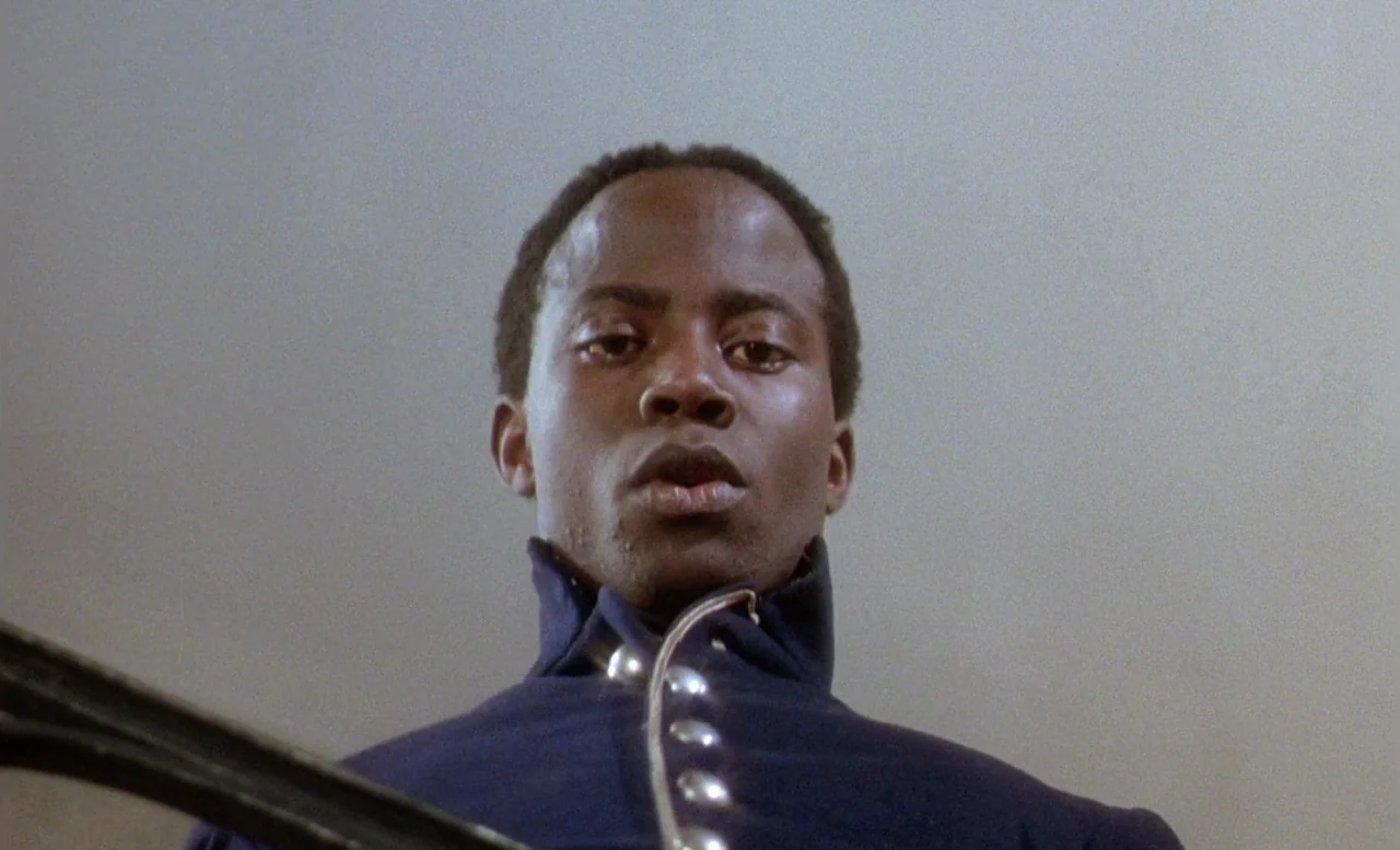 The Beast (1975) movie still: closeup — A man stands on a staircase against a plain, light gray background. He gazes downward directly into the…
