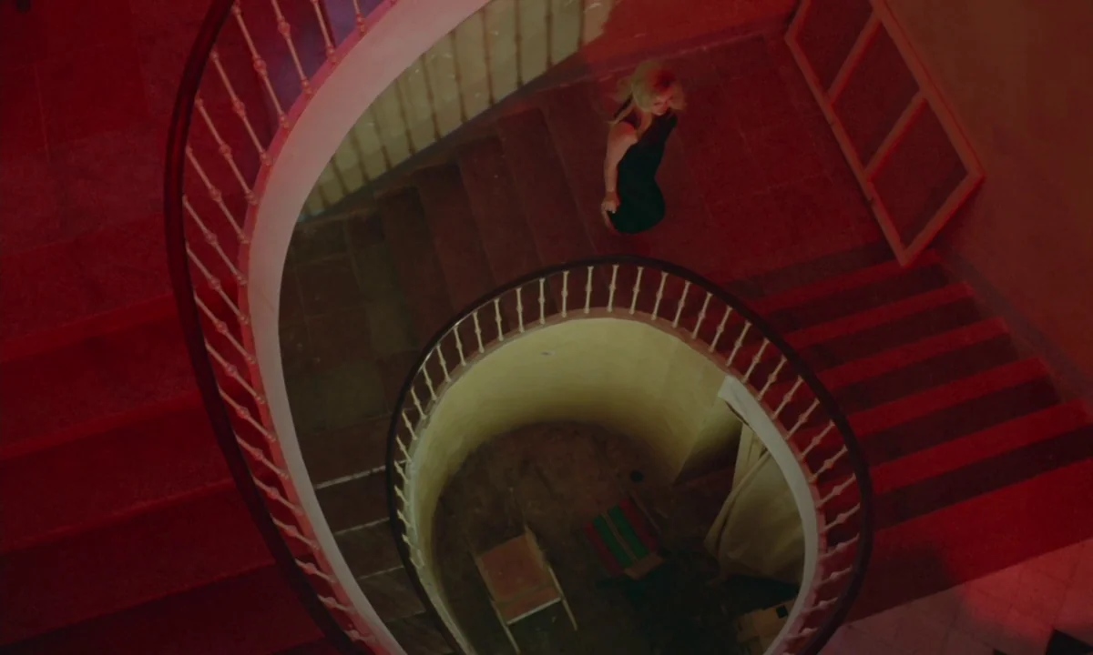 Vampyros Lesbos (1971) movie still: wide — A minimalist circular staircase features a deep red stairwell with a curved banister railing. A woman in…