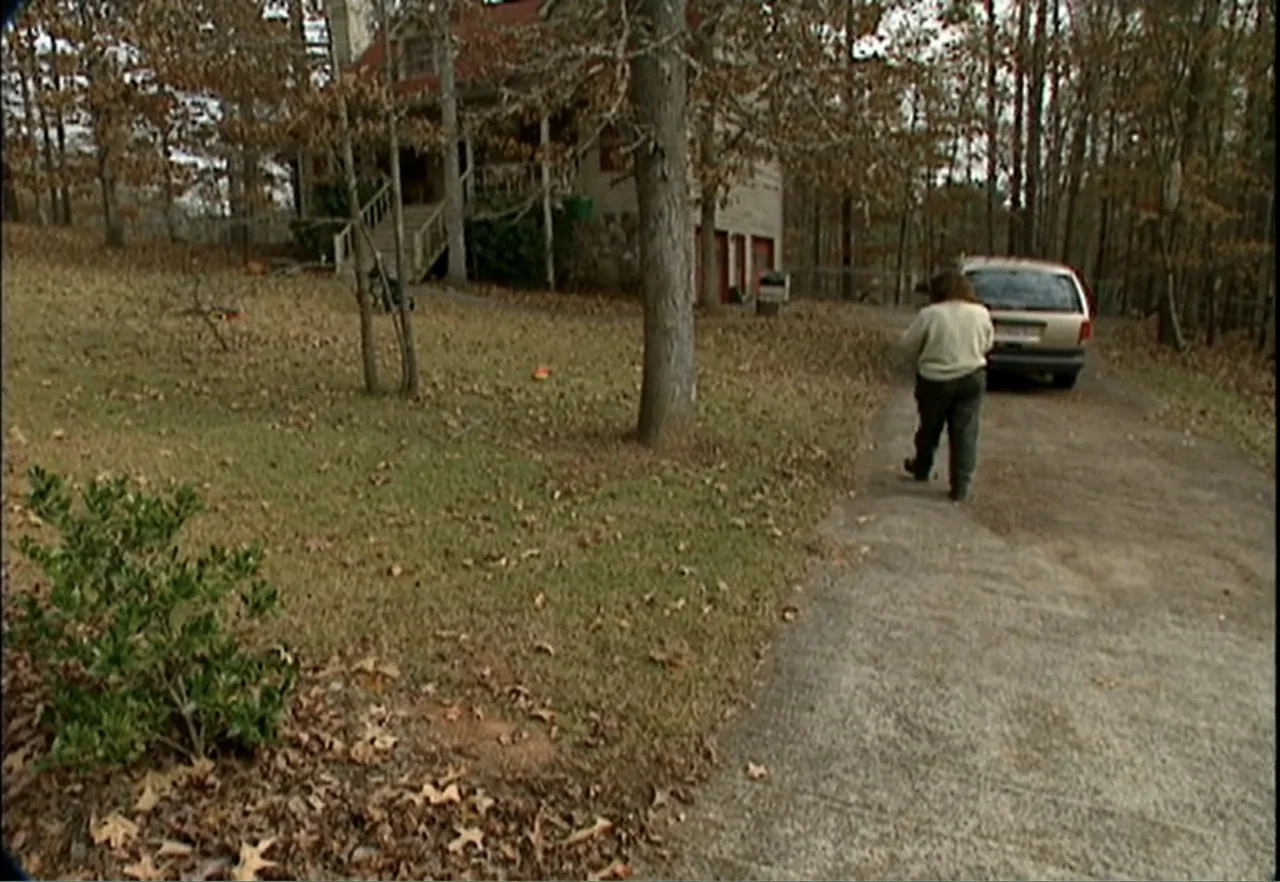Broken Child (2000) movie still: fullbody — A residential driveway leads to a suburban home surrounded by dense, leafless trees. A woman in a beige…