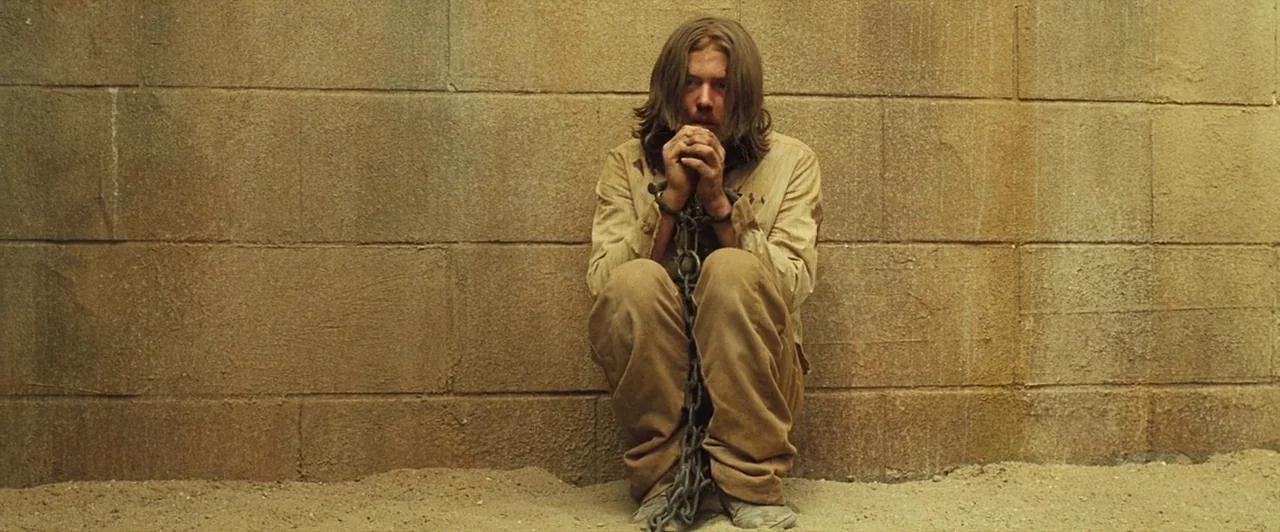 The Proposition (2005) movie still: medium — A man sits on a dirt floor against a rough concrete block wall. He has long brown hair and is wearing…