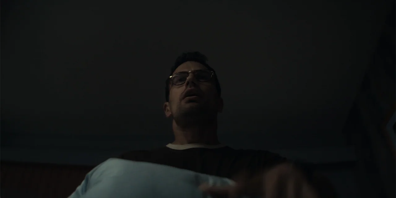 The Monkey (2025) movie still: medium — A dark interior room with muted walls and a soft light source above. A man wearing glasses sits in bed…