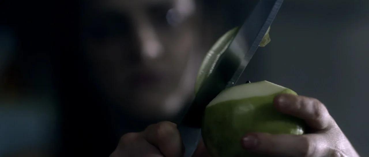 The Lullaby (2019) movie still: closeup — A dark indoor setting focuses on a person preparing food in dim light. The person holds a green pear and…
