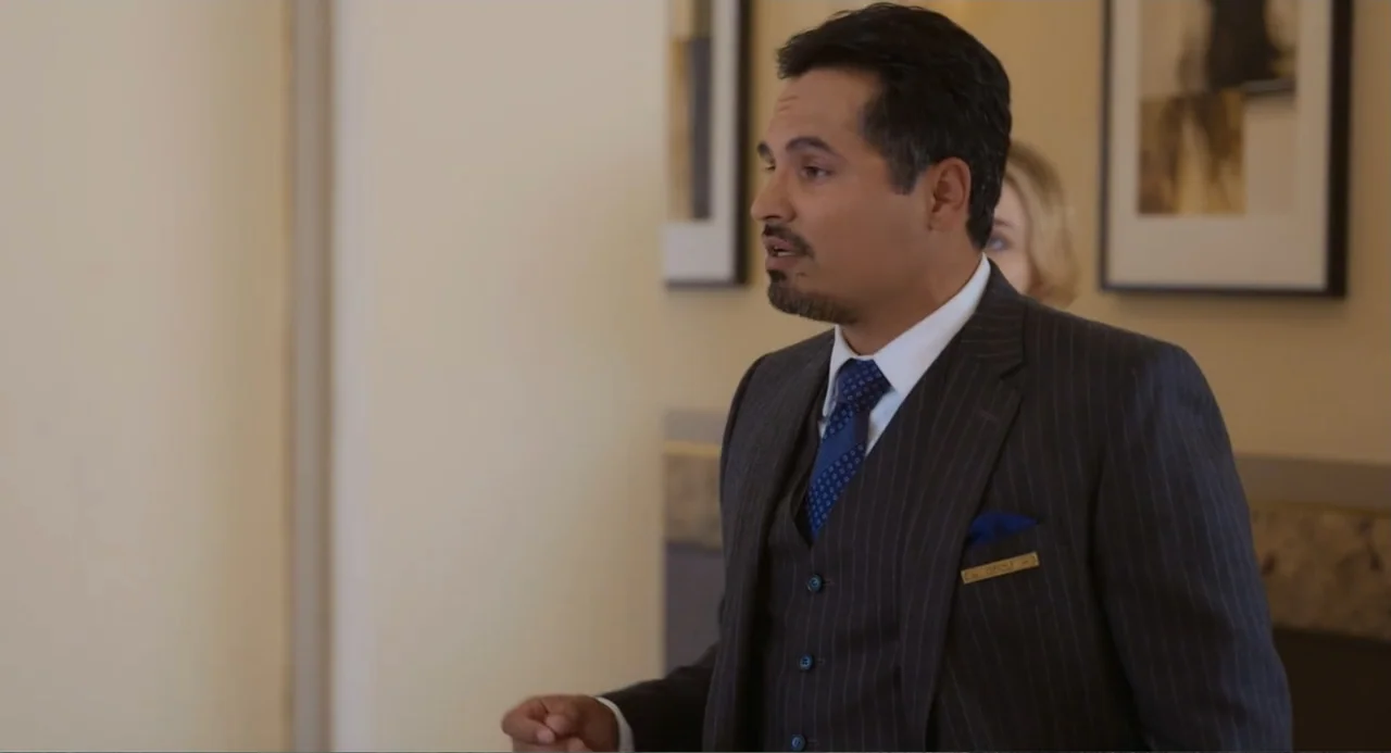 Tom & Jerry (2021) movie still: medium — A man wearing a suit stands in a beige-toned hotel lobby area. He speaks while another person is partially…