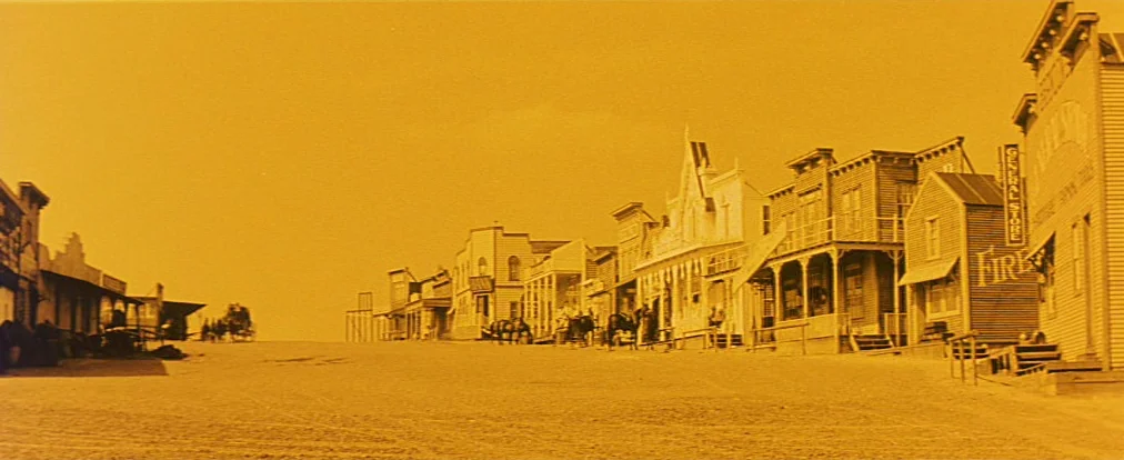 Lemonade Joe (1964) movie still: establishing — A quiet, dusty western town street features wooden buildings with porches and balconies. Several…