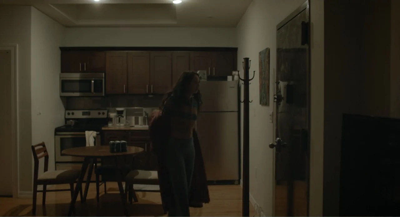 This Closeness (2024) movie still: medium — A residential apartment with a kitchen area and dark wood cabinetry. A woman stands in the middle of the…
