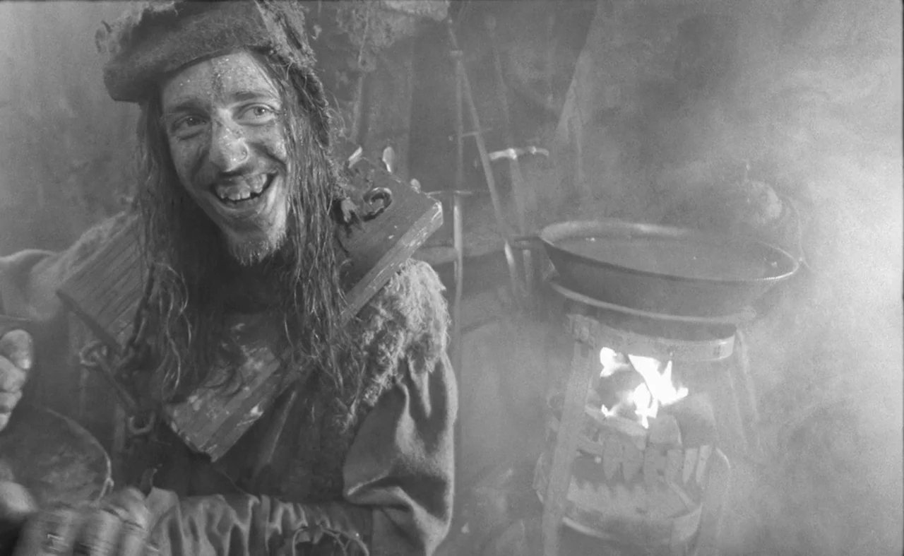 Hard To Be A God (2013) movie still: medium — A dimly lit stone enclosure filled with thick haze and smoke. A man wearing a wooden pillory smiles…