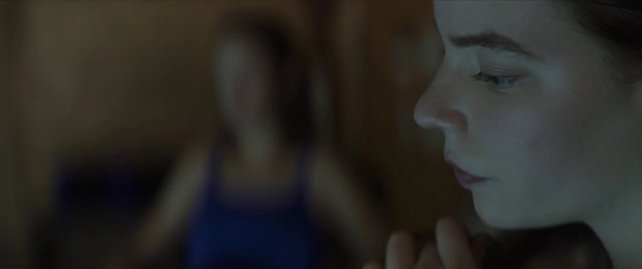 Thoroughbreds (2017) movie still: closeup — A dark indoor setting with a blurred, out-of-focus person sitting in the background. A young woman's…
