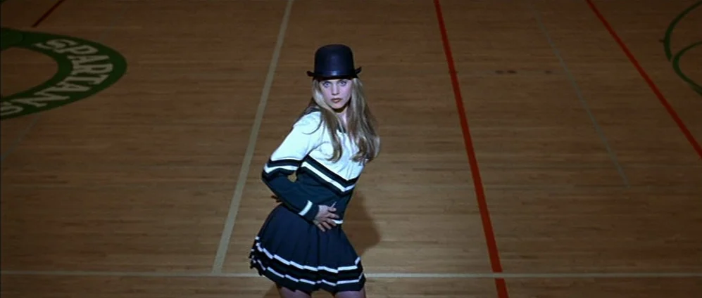 American Beauty (1999) movie still: fullbody — The polished wooden surface of a school basketball court with painted team markings. A young blonde…