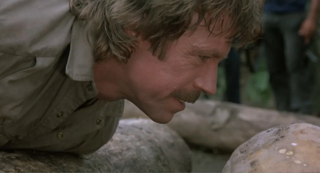 Firewalker (1986) movie still: medium — A man lies on the ground in a dense outdoor natural environment. The man watches a rock while positioned low…