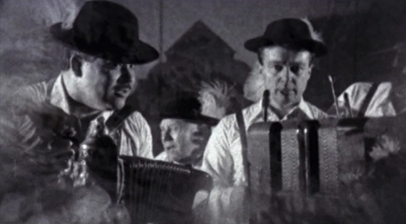 The Saddest Music in the World (2003) movie still: medium — Two men play button accordions in a dim, dark tavern setting. A third man is partially…