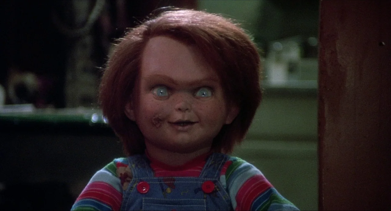 Child’s Play (1988) movie still: closeup — A doll with damaged facial features stands against an indistinct dark interior background. The doll looks…