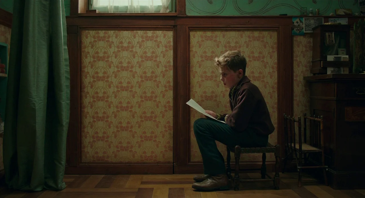 Jojo Rabbit (2019) movie still: medium — A small room features patterned wallpaper above dark wood paneling and a wooden floor. A young boy sits in…