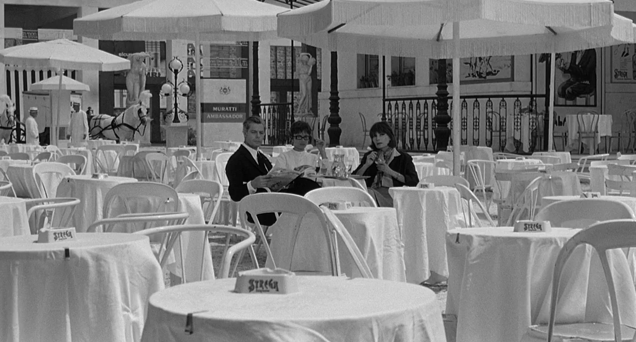 8 1/2 (1963) movie still: wide — An outdoor cafe features numerous white-clothed tables shaded by large umbrellas in a plaza. A man and a woman sit…