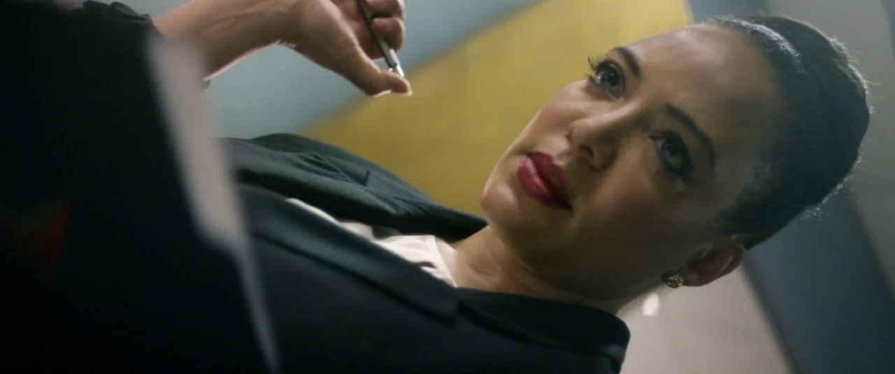 Officer Downe (2016) movie still: closeup — A woman sits in a modern office space with a paneled wall behind her. She holds a silver pen while…